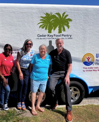 Four people stand in front of a refrigerated trailer with "Cedar Key Food Pantry" written on it. Two palm trees are silhouetted above the text. A United Way North Central Florida logo is visible on the right side of the trailer.