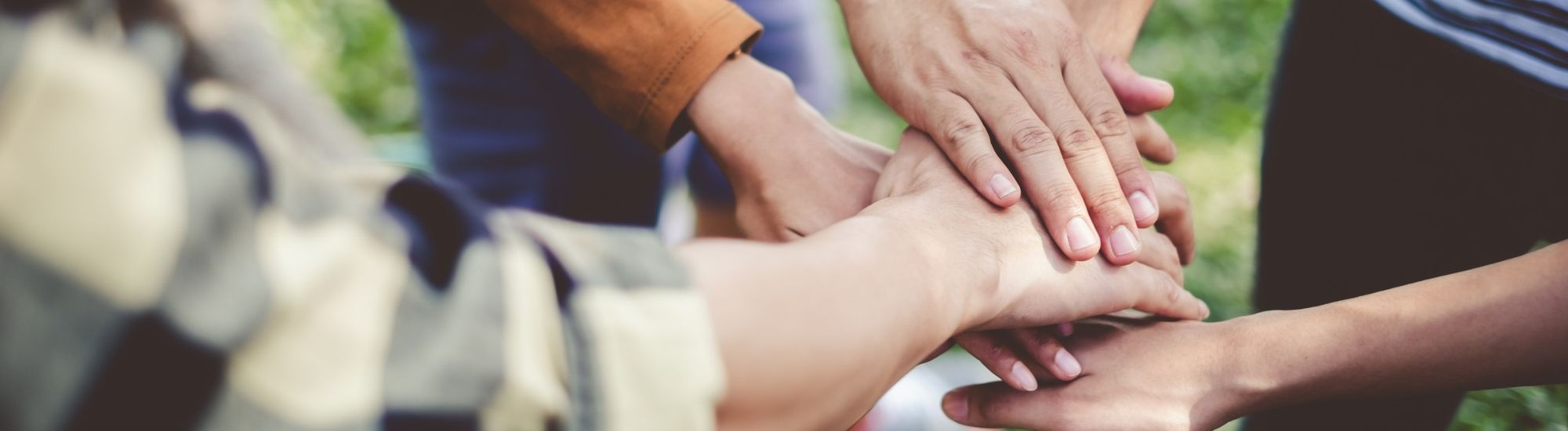 Multiple hands reach in and stack together over a grassy outdoor area, symbolizing teamwork and support.