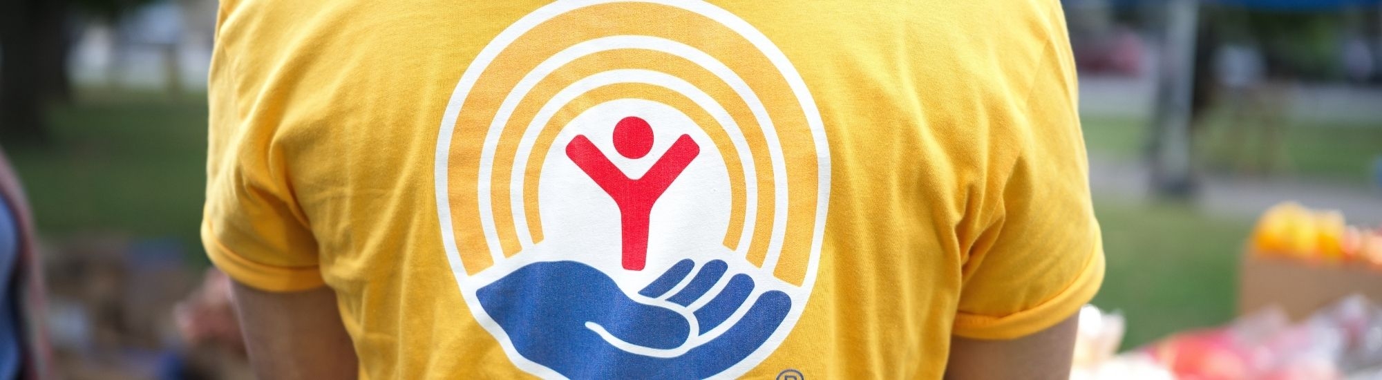 Back view of a person wearing a yellow T-shirt with a circular rainbow logo and a red hand symbol on the center, outdoors with blurred greenery in the background.