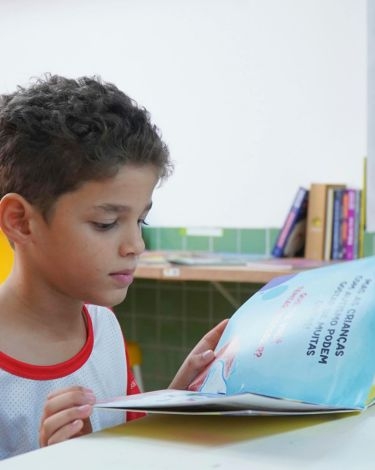 A child with curly hair reads a book with illustrations and text on a bright blue page.