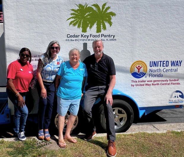 Four people stand in front of a trailer with "Cedar Key Food Pantry" and "United Way North Central Florida" logos. The trailer is a food delivery vehicle.