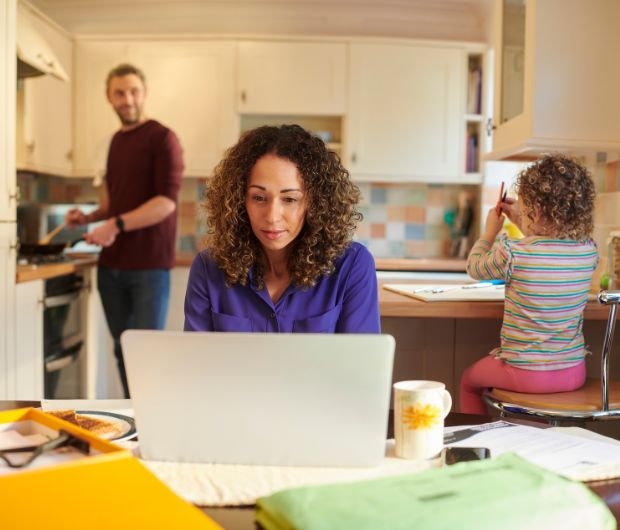 A woman works on a laptop in a kitchen while a child draws nearby and a man cooks in the background.