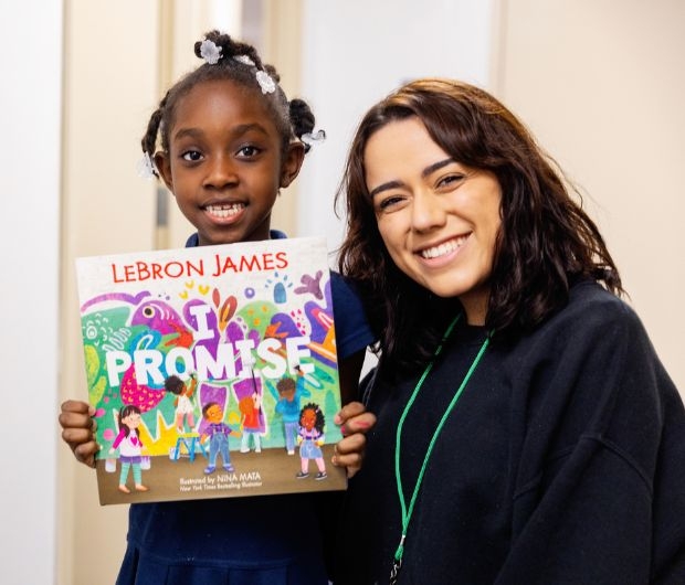 A child holds up the book "I Promise" by LeBron James, standing next to an adult who is also smiling at the camera.