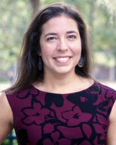 A woman with long dark hair smiles at the camera, wearing a dark purple dress with a floral pattern and decorative earrings.